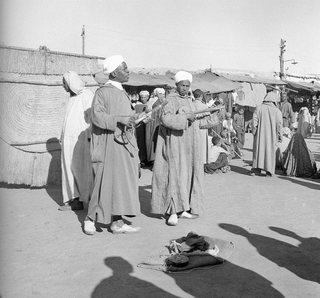Two Moroccan street musicians play in a crowded street; in recent times in Morocco some women have started to assert themselves in order to reach a modern state of emancipation and to free themselves from their condition of slavery to men; on the contrary other women have not felt the need of this cultural revolution and have kept on respecting the atavistic habits. Marrakech (Morocco), December 1954.